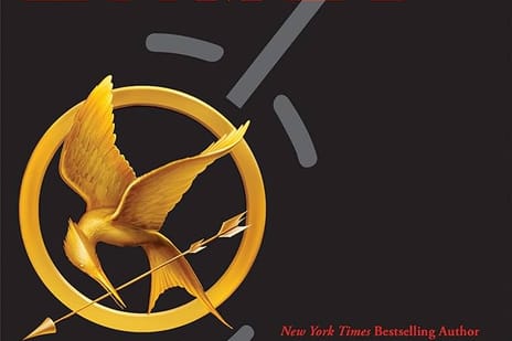 A black background with a gold pin on it. The pin has a mockingjay holding an arrow encircled by an elongated bow.